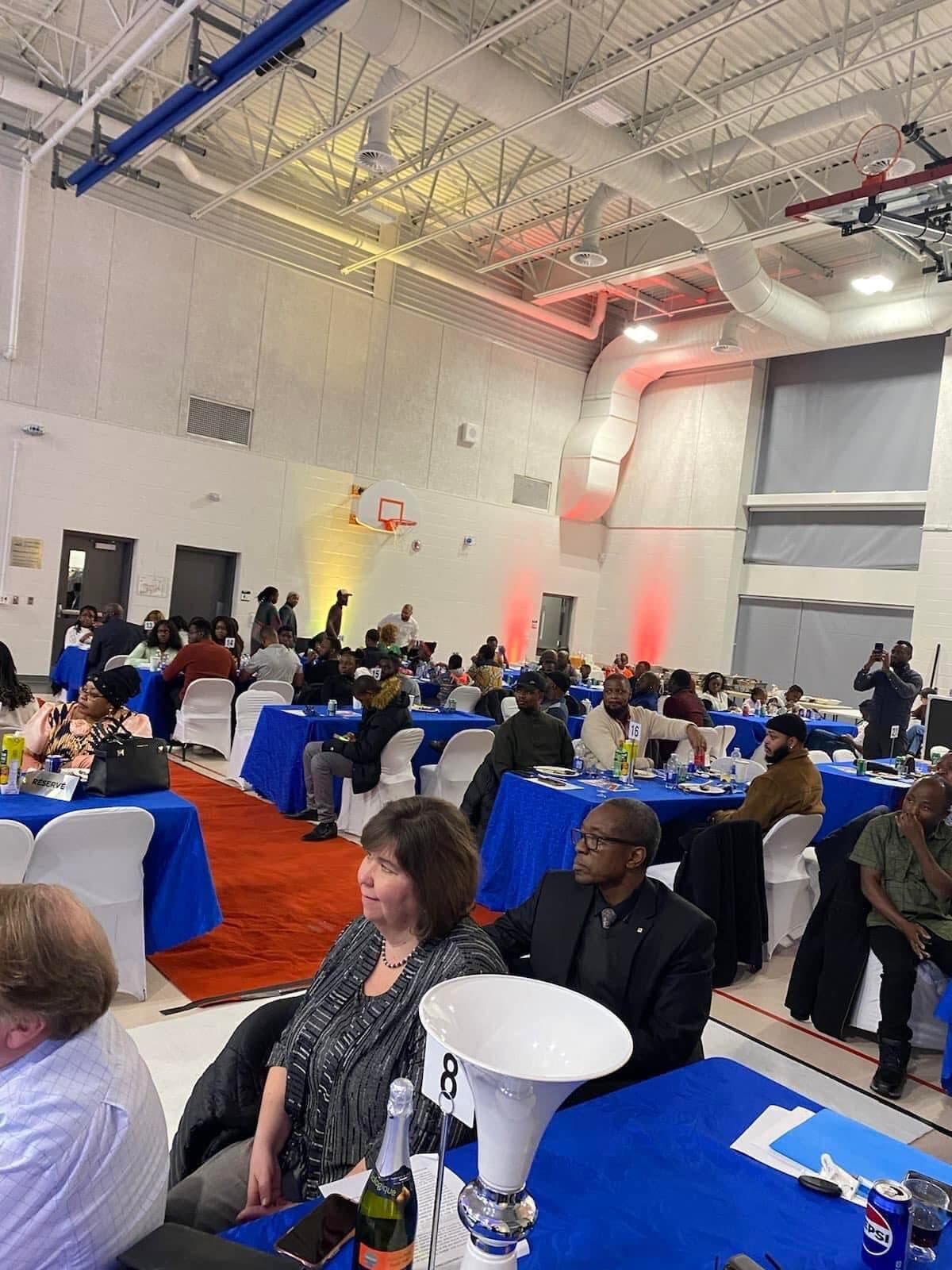 Une salle de gala dans un gymnase : des invités assis à des tables aux nappes bleues, de part et d’autre d’un tapis rouge.