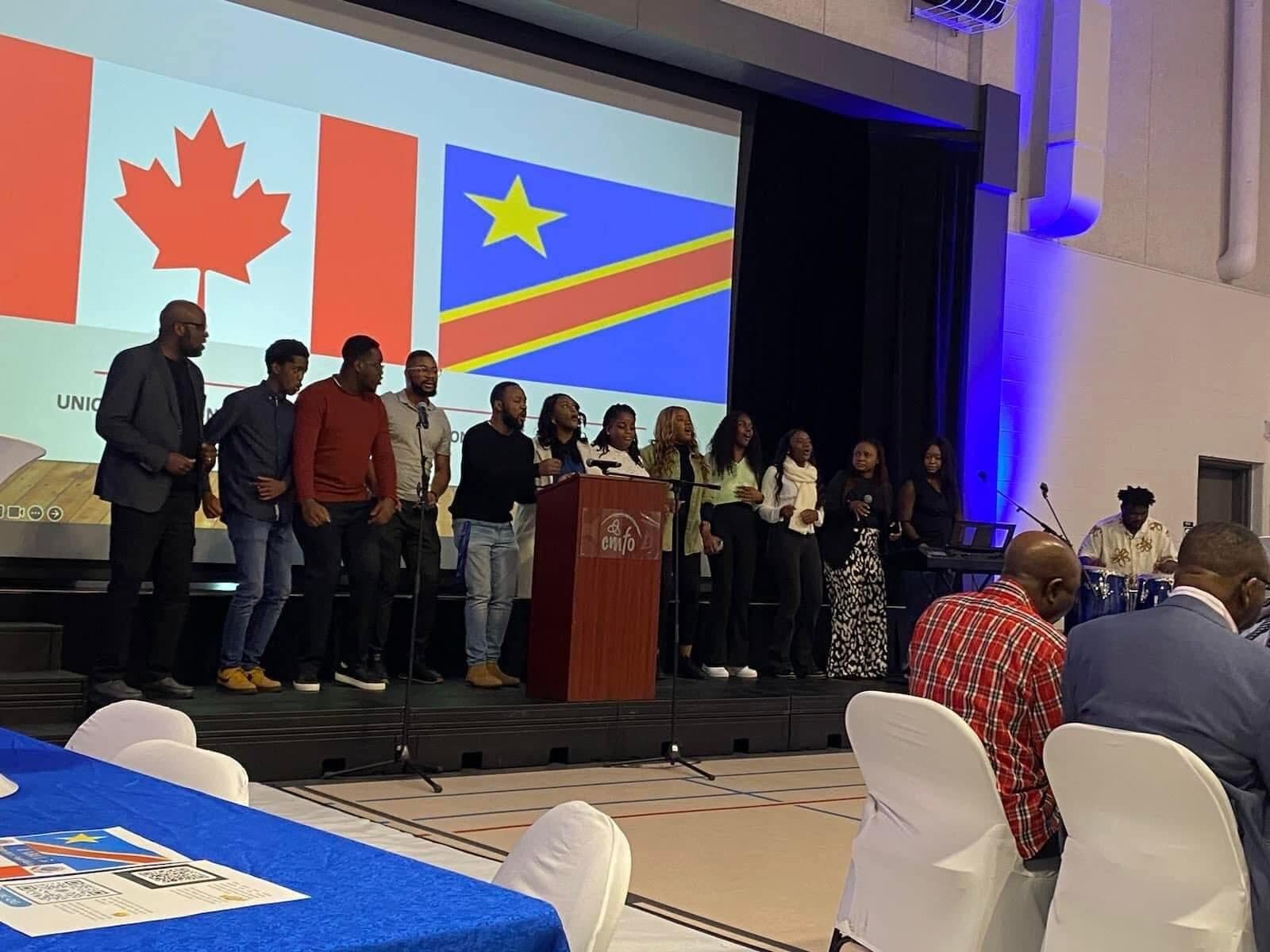 Sur scène, un groupe chante devant un écran aux drapeaux du Canada et de la République démocratique du Congo ; des invités sont assis aux tables.