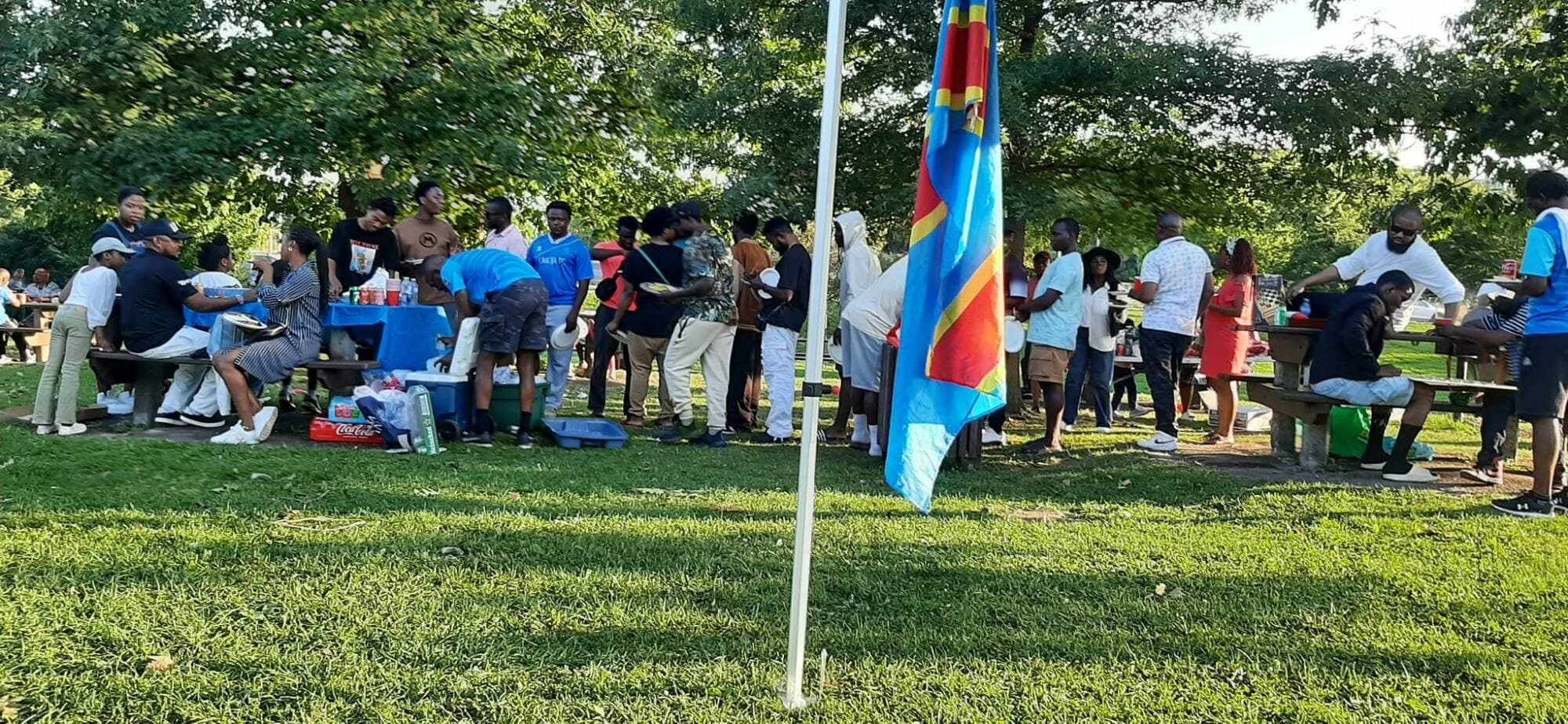Pique-nique dans un parc : des dizaines de personnes autour de tables, sous les arbres ; au premier plan, un drapeau de la République démocratique du Congo.